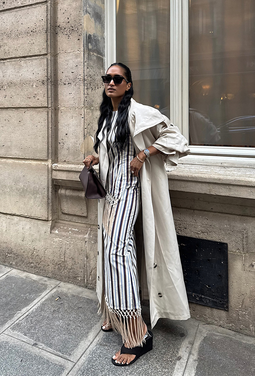 The sandal trend wedge flip-flops are shown in a photo of a woman standing outside wearing a tan trench coat over a striped top and a matching fringe maxi skirt styled with black flip-flop wedges, a black clutch, and black sunglasses