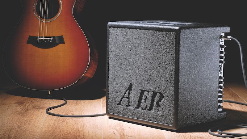 An AER acoustic amp on a wooden floor with an acoustic guitar in the background