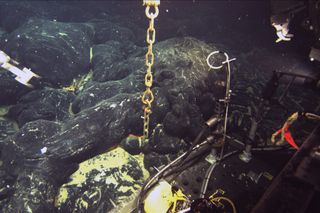 A chain still attached to an ocean-bottom hydrophone (OBH) comes directly out of the seafloor at Axial Seamount where a lava flow erupted in 2011 has buried the instrument to a depth of about 1.5 meters (5 feet). The front of the Jason remotely operated vehicle is in the lower right and its manipulator arms are visible in the upper left and right.