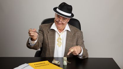 A stereotypical con artist holds a pen and points at a contract on his desk.