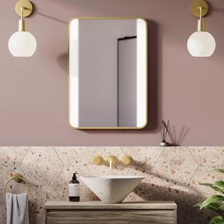 brass edged mirror with LED strips on either side on pink wall, with wall mounted brass and white globe wall lights either side, terrazzo tiles below and sink