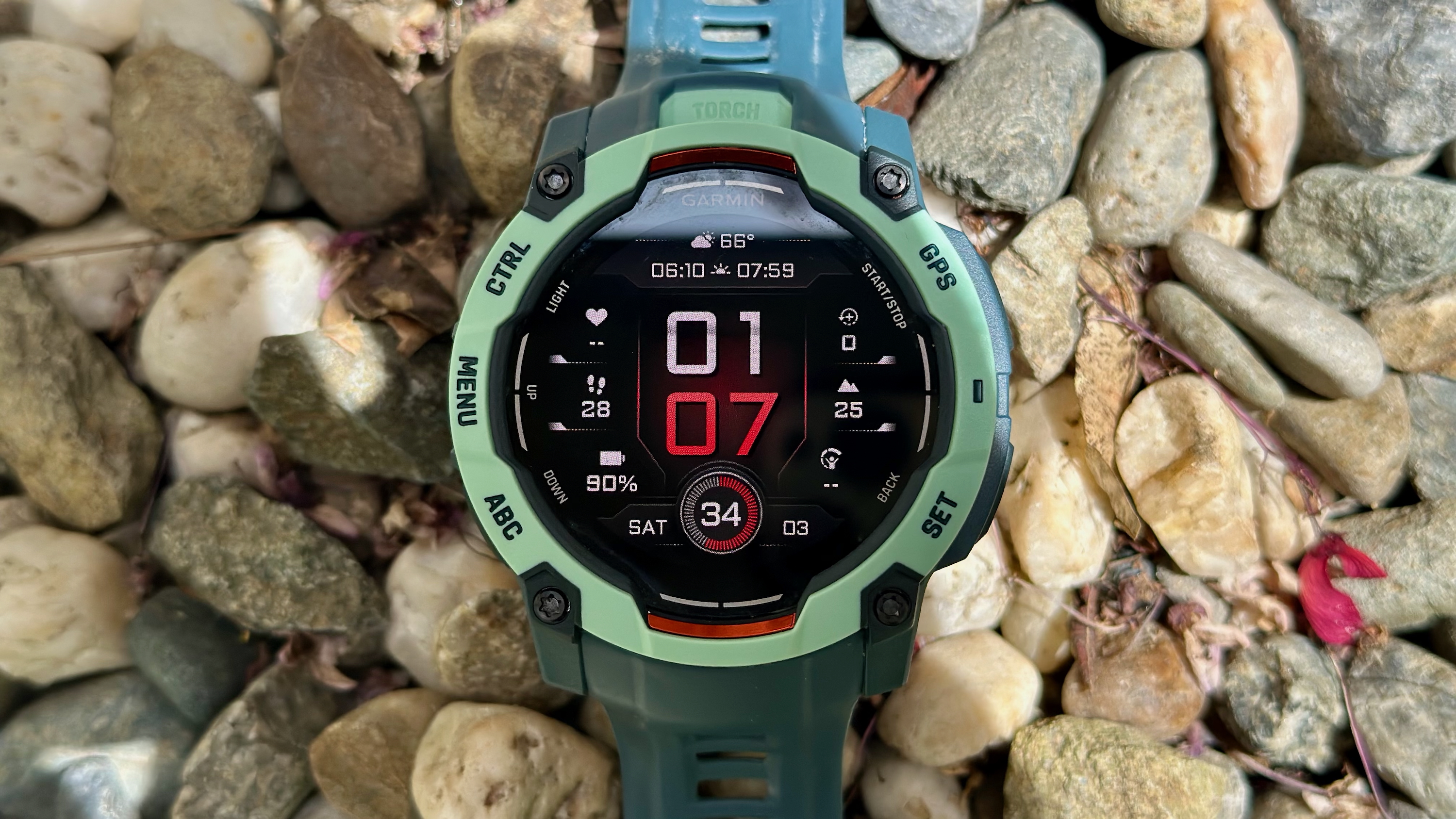 The Garmin Instinct 3 50mm AMOLED sitting atop pebbles and small rocks, the red-and-white watch face showing various data like the time & date, weather, elevation, remaining battery life, and VO2 Max.