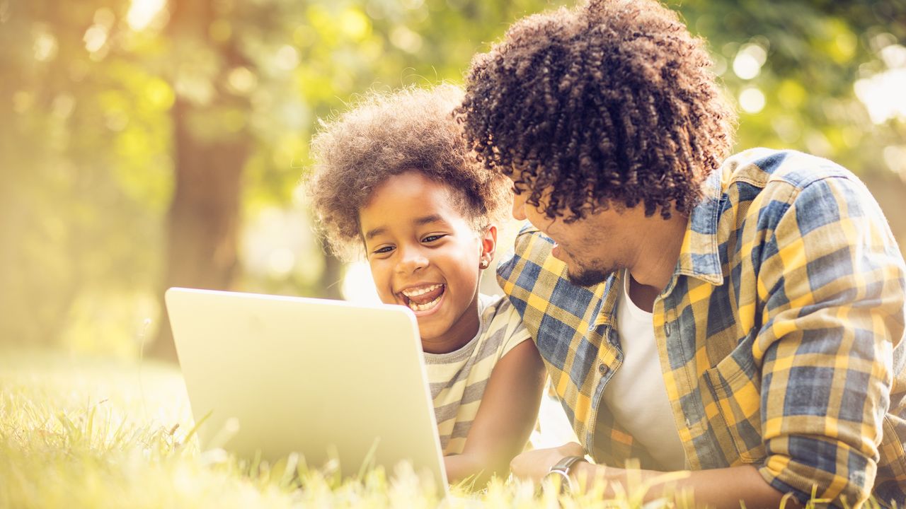 A dad and his daughter laugh and smile while looking at a laptop together in the park.