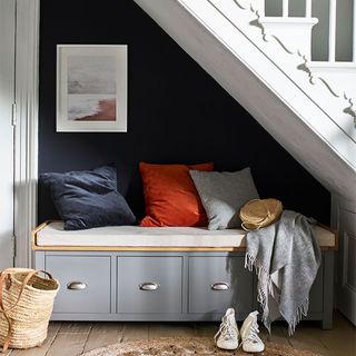 an under stairs seating area with a grey storage bench seat with dark blue, orange and grey cushions on top, set against a dark blue wall