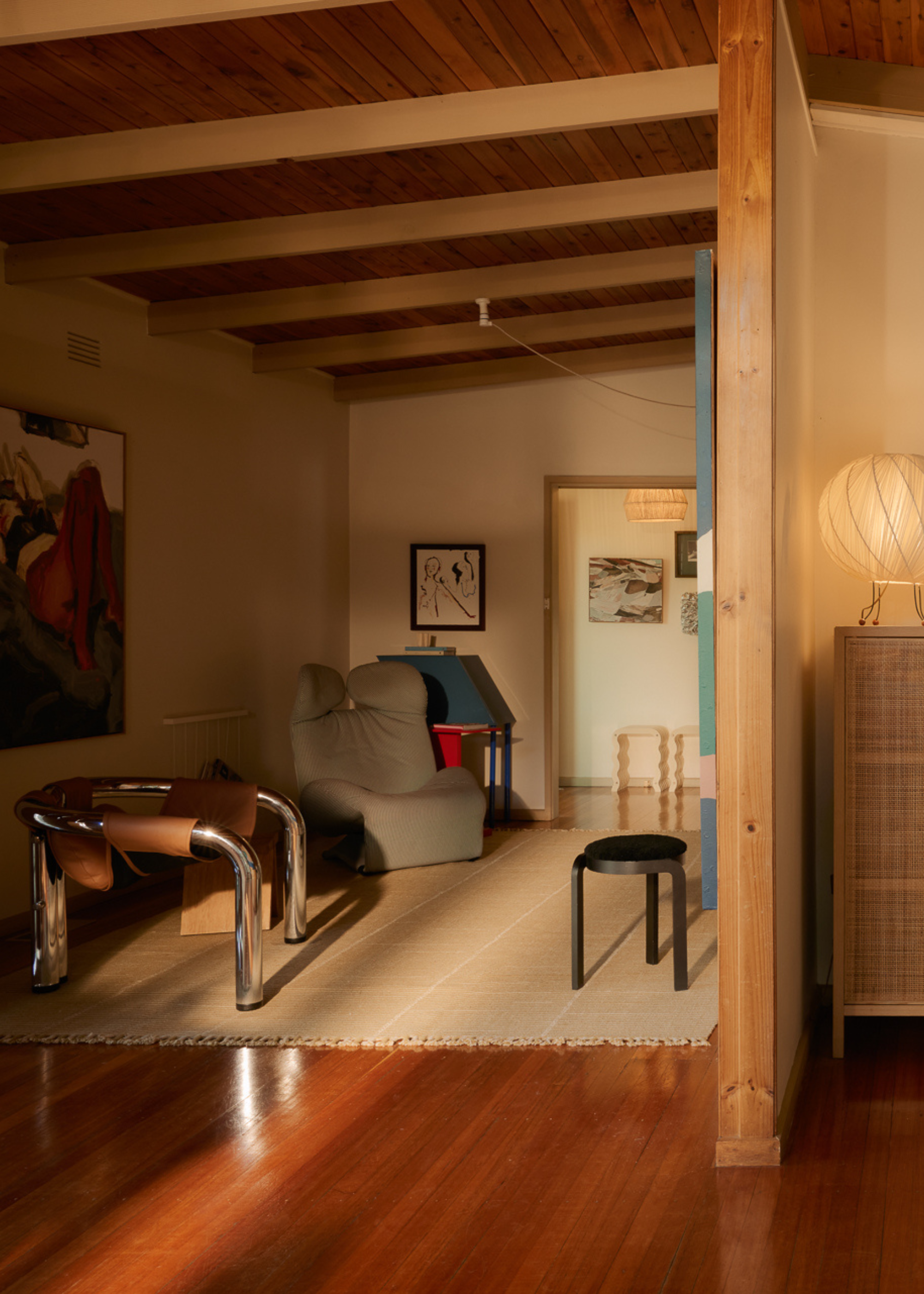 A warm living room with exposed beams and wood panel ceilings, a metal tube chair with a leather sling, a gray recliner, a black stool, a painting, a sideboard, and a paper lamp