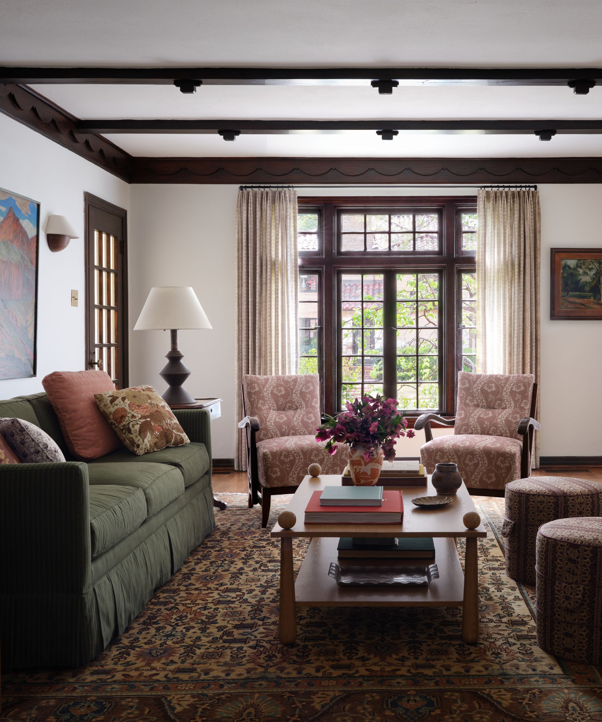 A traditional living room with a green pinstripe sofa, patterned armchairs, and dark wood ceiling beams featuring decorative scalloped trim