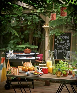 Outdoor dining ideas with a Bonfire Night party sign on a blackboard, barbecue and sticky apples and drinks on a table, with lanterns hanging from foliage canopy