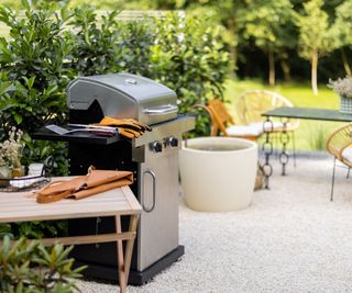 A stainless steel grill in a back yard with gravel, grass and a table