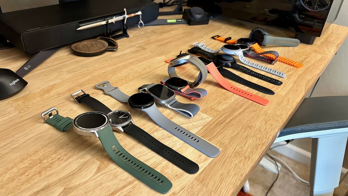 A side view of thirteen smartwatches sitting side-by-side on a desk. In order, they are the OnePlus Watch 2R, Withings ScanWatch 2, Google Pixel Watch 3, Apple Watch Ultra 2, Fitbit Sense, Samsung Galaxy Watch 5 Pro, Fitbit Charge 6, Samsung Galaxy Watch 6 Classic, Garmin Fenix 8, Coros Pace Pro, Amazfit Cheetah Pro, Samsung Galaxy Watch Ultra, and Garmin Instinct 2X Solar.