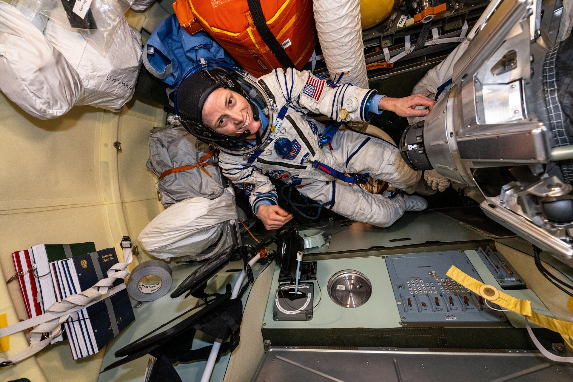 a woman in a white space suit, but no helmet, floats in a space station module surrounded by equipment. The walls are tan.