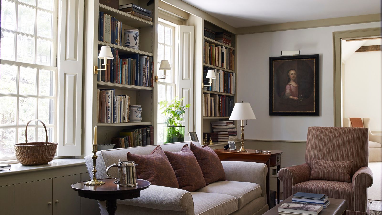 A neutral living room, home library space with plush sofa, chair, bookshelves, and large bright windows with white shutters.