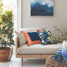 A living room with a sofa with a wooden frame, an abstract artwork hanging on the back wall and a potted tree placed next to the sofa