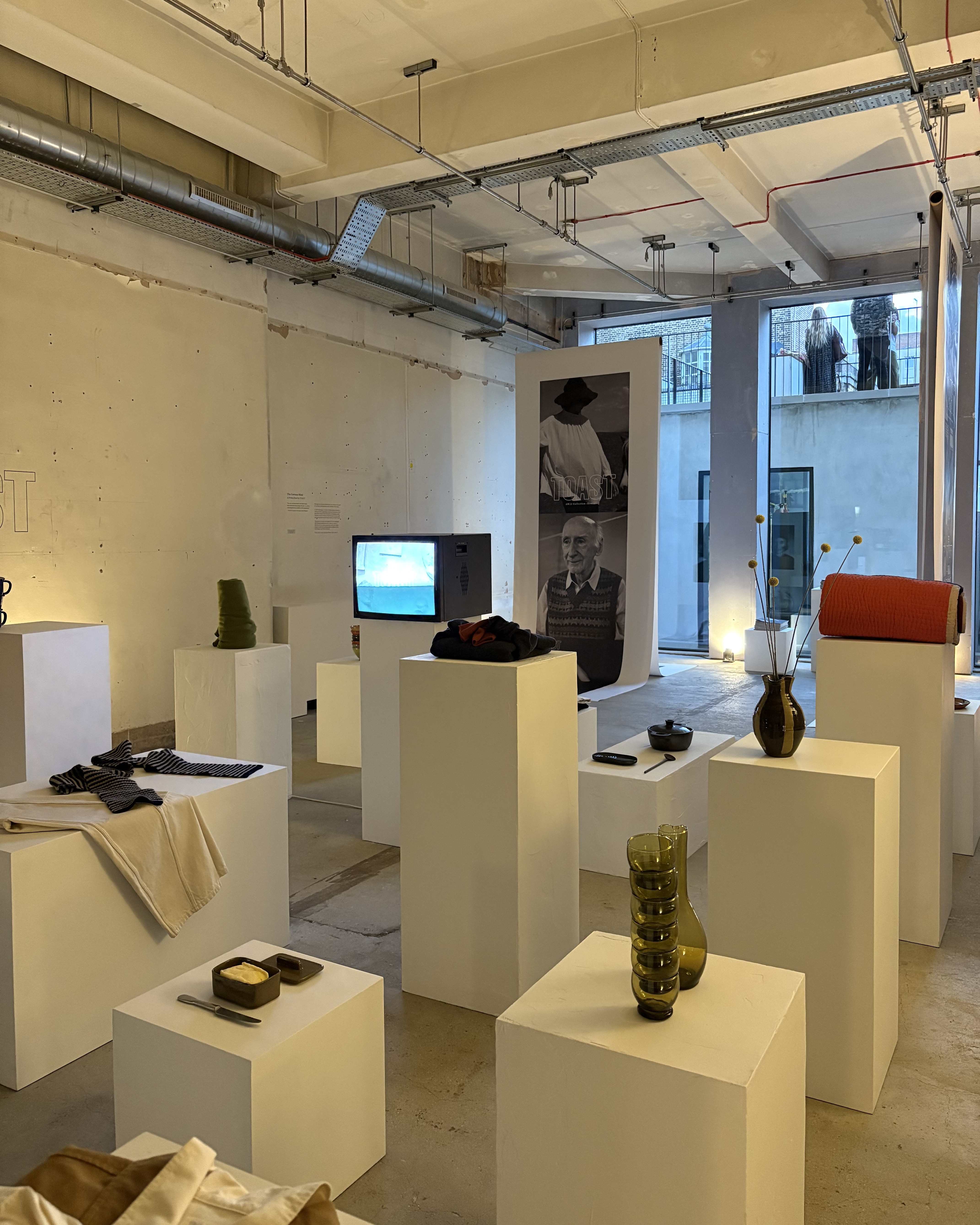 Installation view of a fashion and design exhibition featuring clothes, homeware, retro-looking tvs showcasing audiovisual works, large posters, and more.