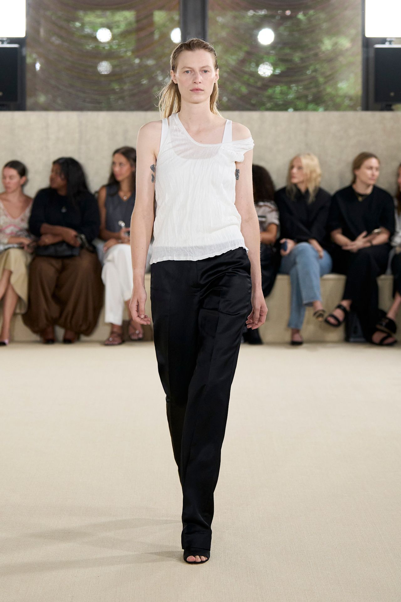 Layering staples for spring 2026 are shown in a photo from Toteme's runway show, with a model walking while wearing a sheer white tank top layered on top of a white tank top styled with black wide-leg trousers and black open-toe mules