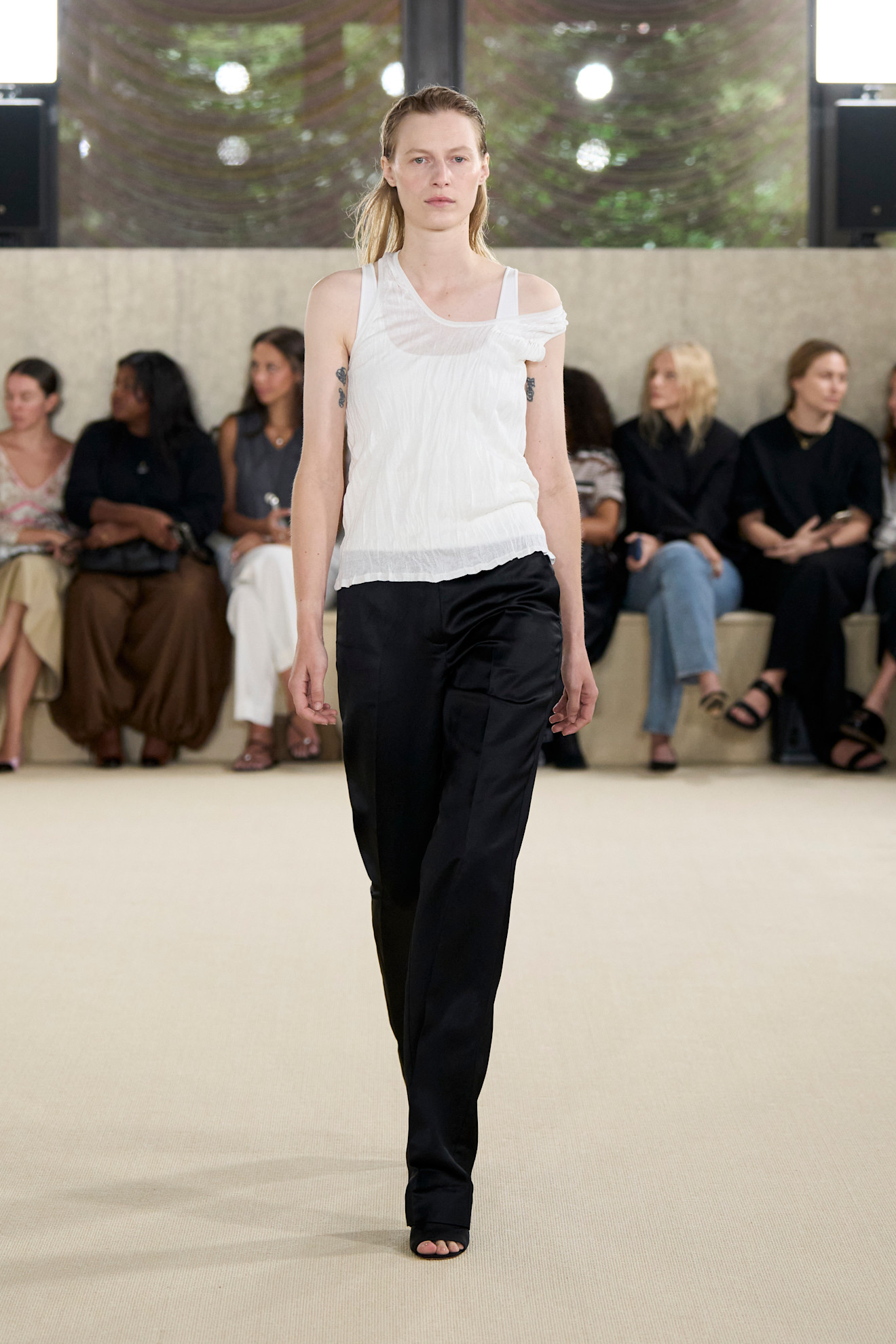 Layering staples for spring 2026 are shown in a photo from Toteme's runway show, with a model walking while wearing a sheer white tank top layered on top of a white tank top styled with black wide-leg trousers and black open-toe mules 