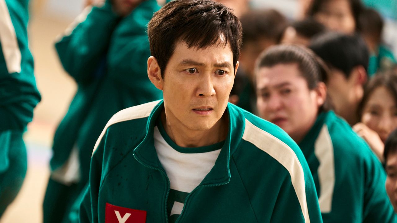 A man with a worried expression (Lee Jung-jae as Gi-hun) stands among a crowd of people in identical green tracksuits, in &#039;Squid Game&#039; season 2.