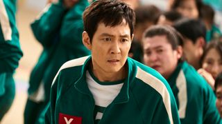 A man with a worried expression (Lee Jung-jae as Gi-hun) stands among a crowd of people in identical green tracksuits, in 'Squid Game' season 2.