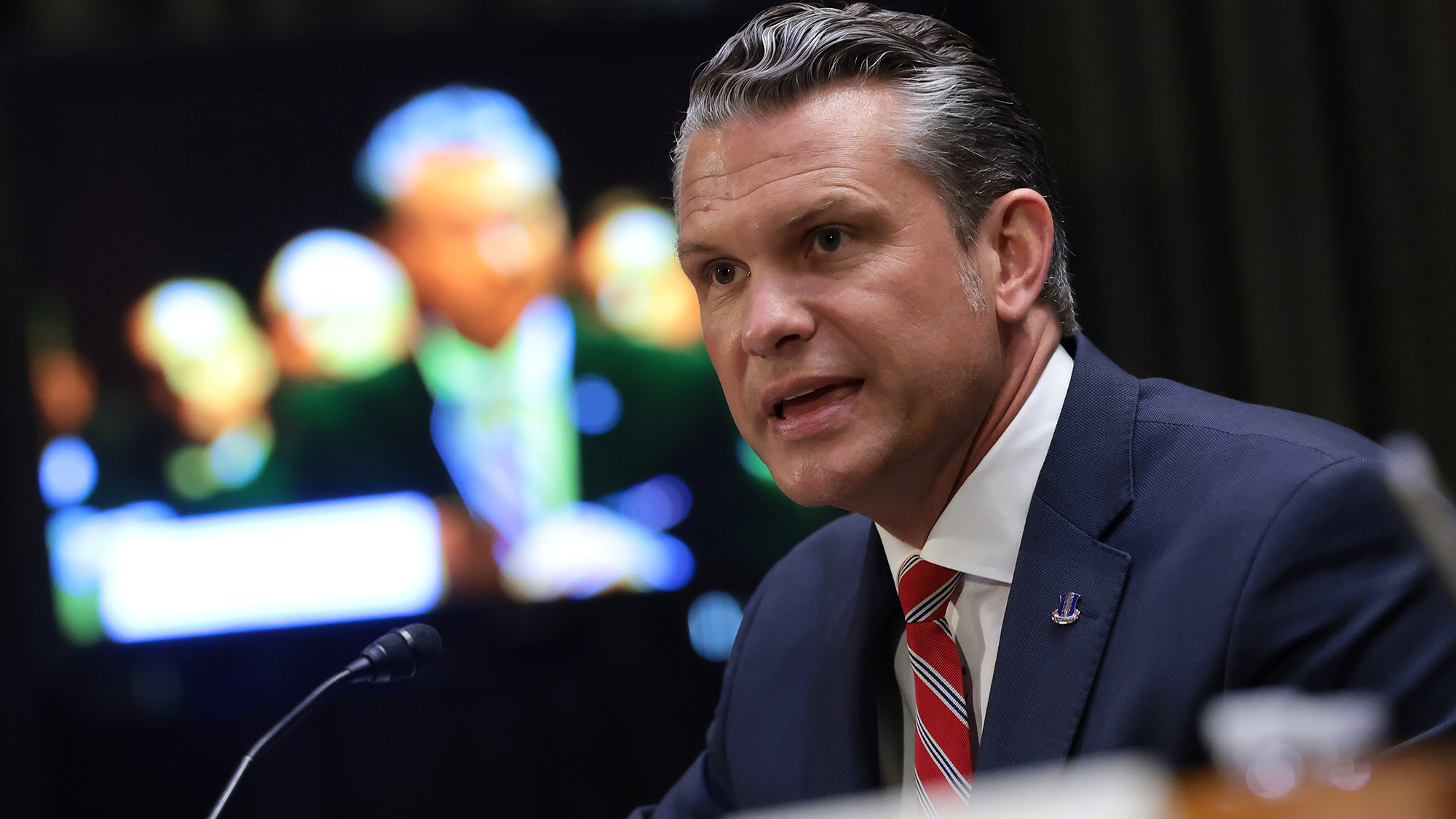 A man with gray hair wearing a navy suit and red tie yells into a microphone