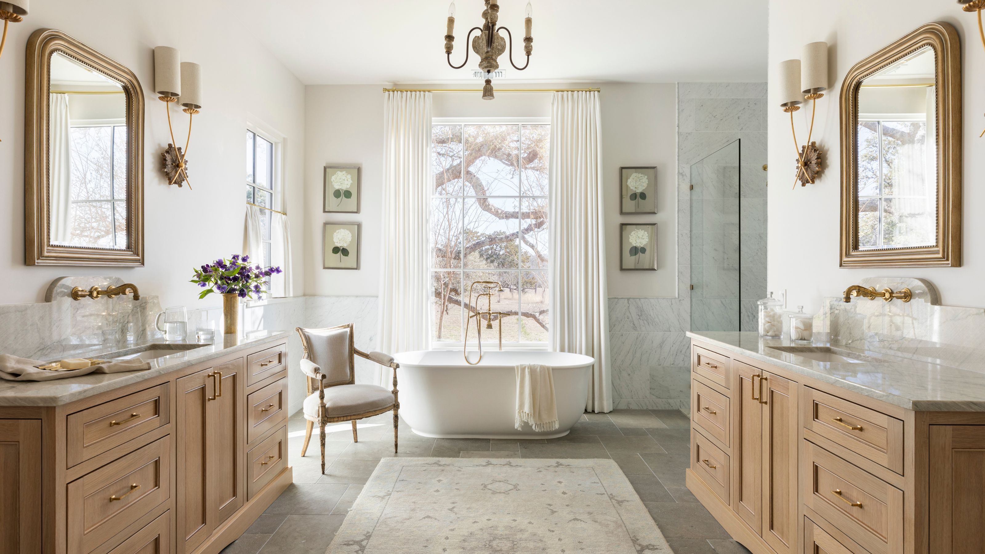 a neutral farmhouse style large bathroom with two oak vanity units and a freestanding tub in front of a window