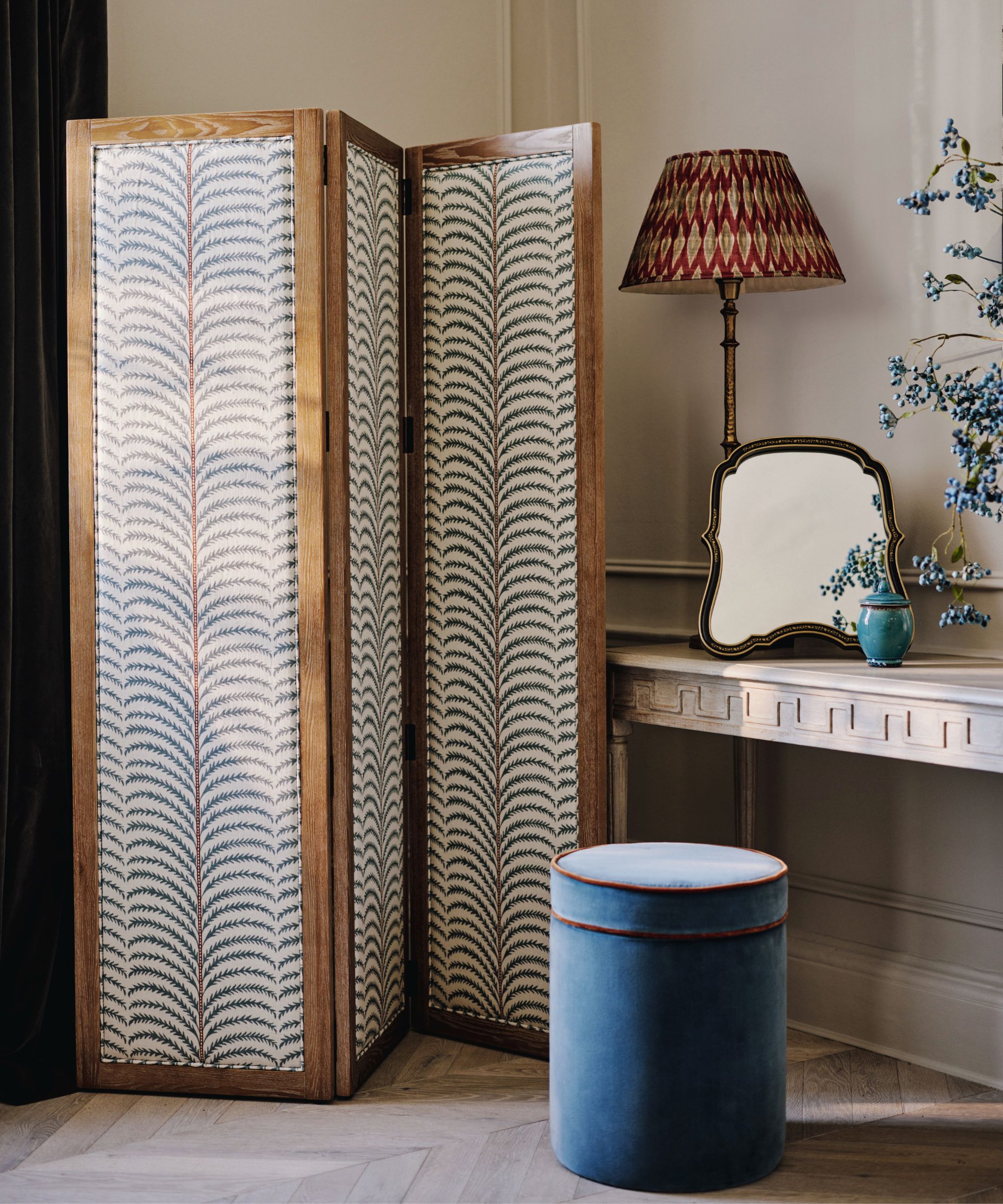 Freestanding folding screen with patterned fabric panels and wood frame, creating a flexible bedroom divider beside a dressing table.
