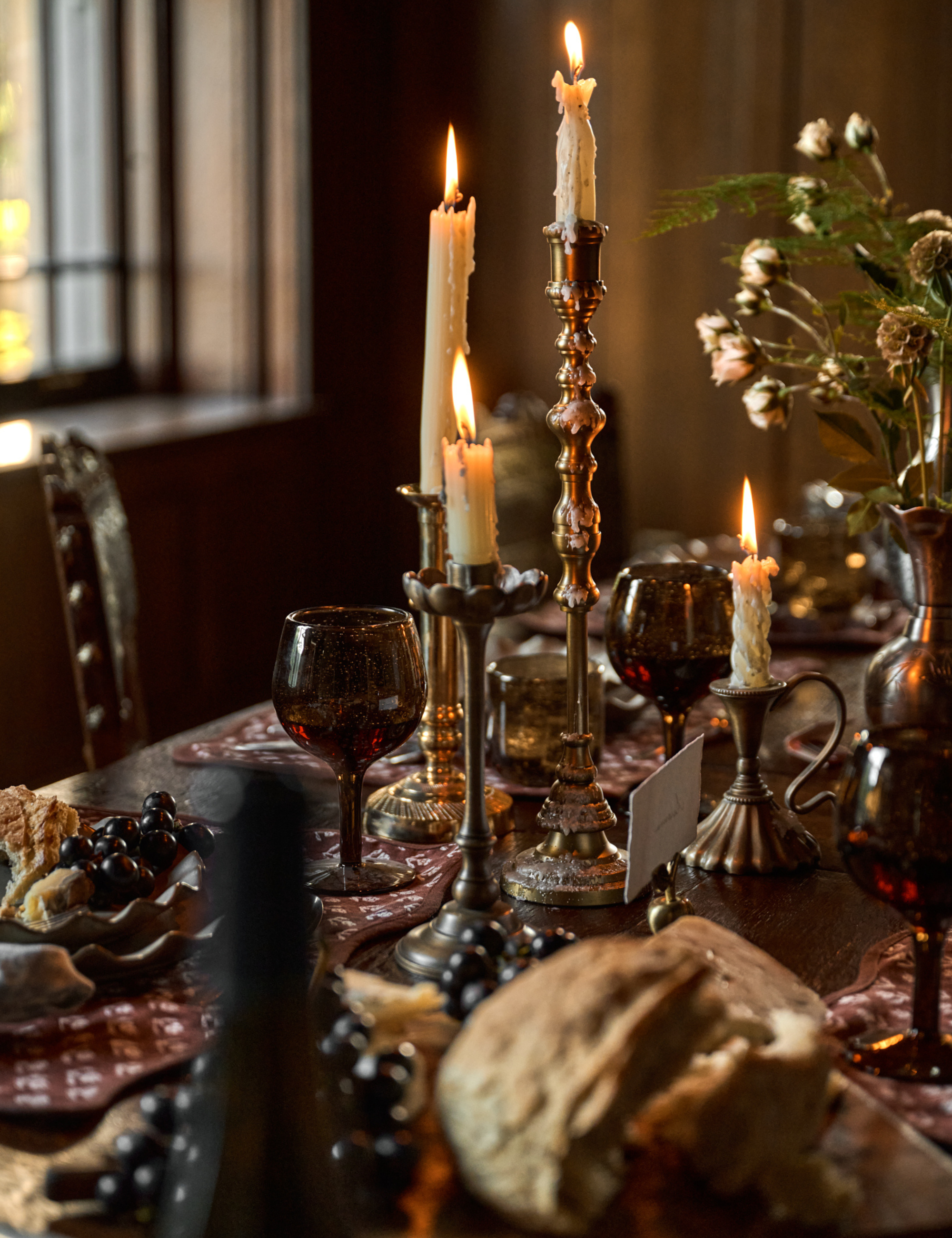 Moody fall tablescape featuring oxblood glassware, lit taper candles set inside antique-inspired candlesticks, and a coordinating patterned table runner.