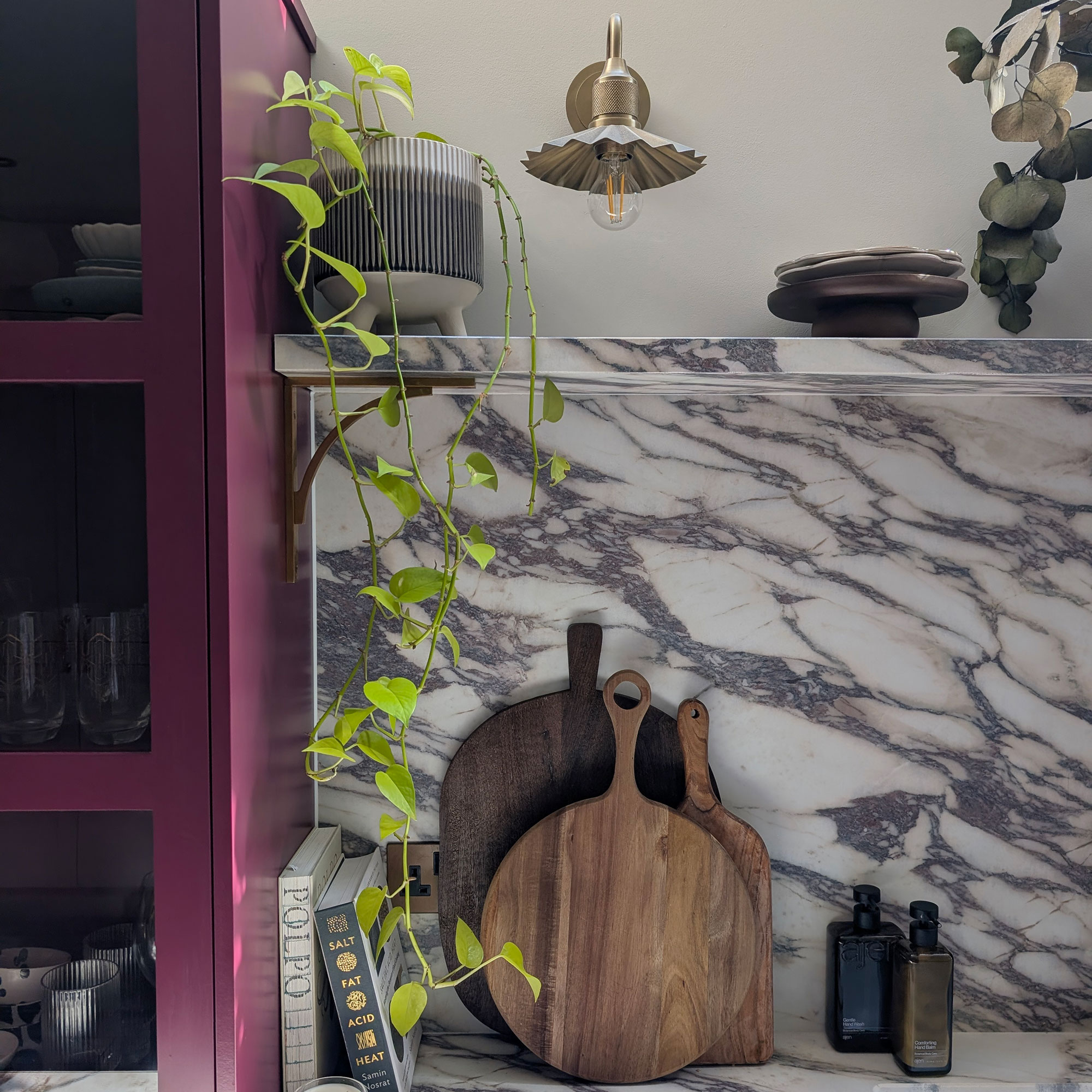 Marble kitchen splashback with round wooden boards leading against it, topped with shelf holding trailing plant with fluted brass wall light behind