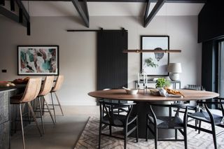 Kitchen-diner with beige walls, large format beige floor tiles, round black and walnut dining table, black wishbone chairs, bar pendant light over table, and brown leather bar stools against a kitchen island