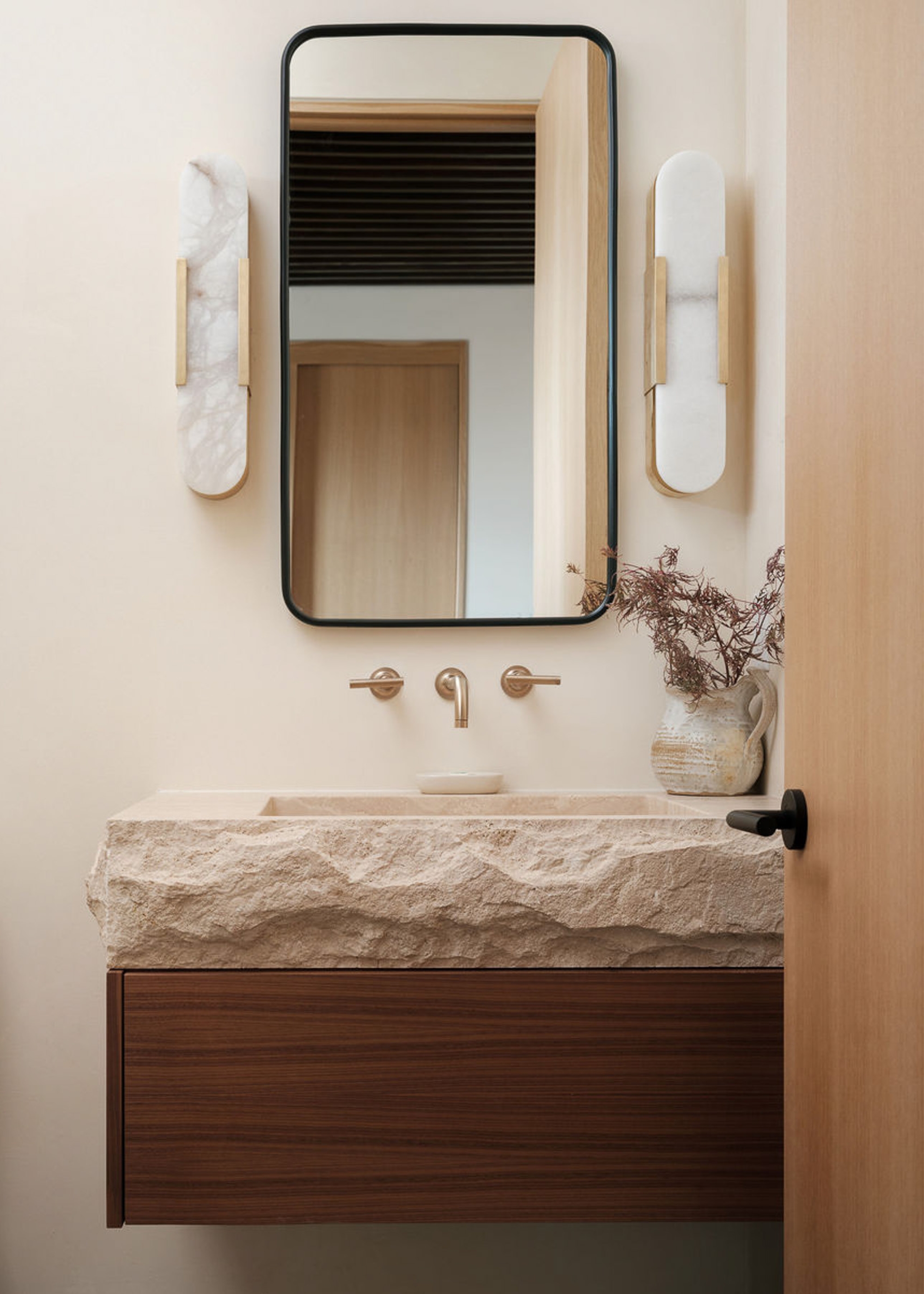 A small bathroom with a floating sink with a raw stone sink and a wooden drawer, matching sconces, a curved mirror, and a vase of stems