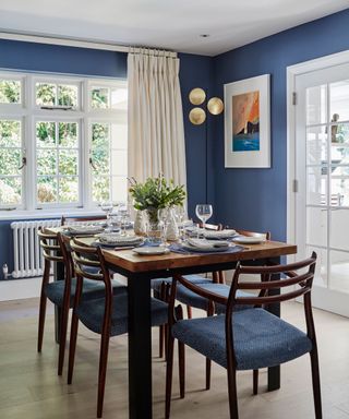 Blue painted dining room, light wooden flooring, large rectangular dining table in dark wood with matching dining chairs, chairs with blue fabric seat upholstery, cream curtains, black and gold rounded floor lamp design, artwork on walls