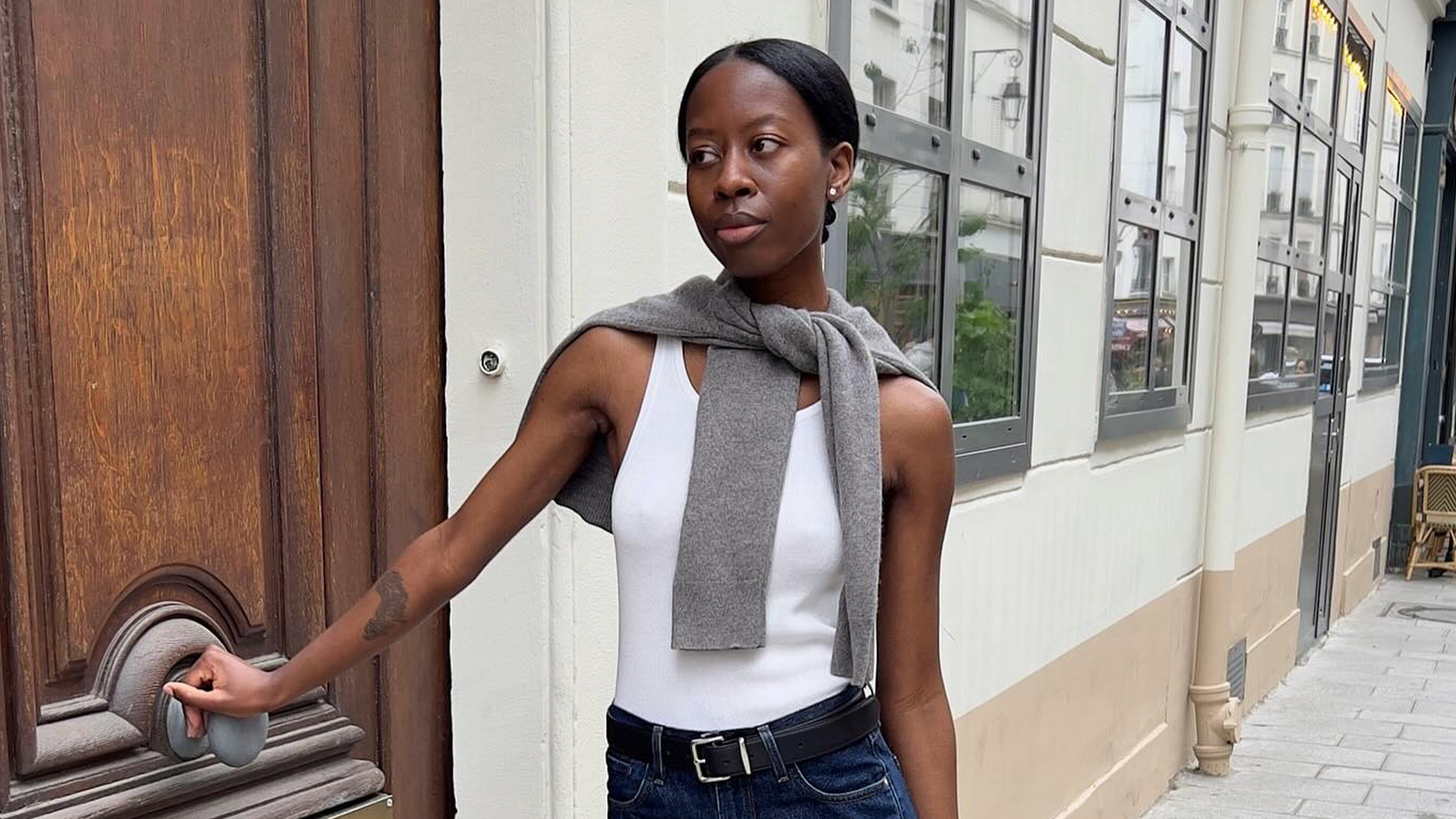 Influencer wearing white tank, grey sweater over shoulders, black belt, and blue jeans standing on the streets of pairs.