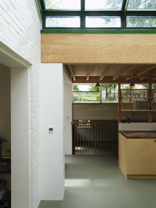 Interior of north London home extension of a Walter Segal house by Hayatsu Architects , grey floor, white brick walls, wooden worktops and shelving, narrow wooden beam low ceiling, black shade ceiling lights, wooden beam under green skylight, window with view of surrounding garden