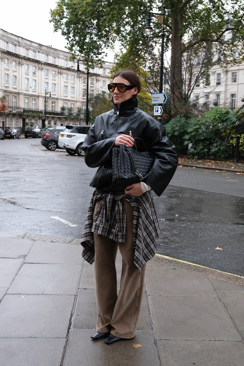 Trendy basics at Nordstrom are shown in a street style photo of a woman standing on the sidewalk in London wearing oversized two-tone aviator sunglasses, a black leather funnel neck bomber jacket styled with a black Bottega bag, a black and brown flannel shirt tied around the waist, brown wide-leg trousers, and black pointed-toe ankle boots.