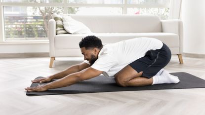 man in a white tshirt and black shorts in a child's pose position on a black yoga mat with a white sofa behind him.