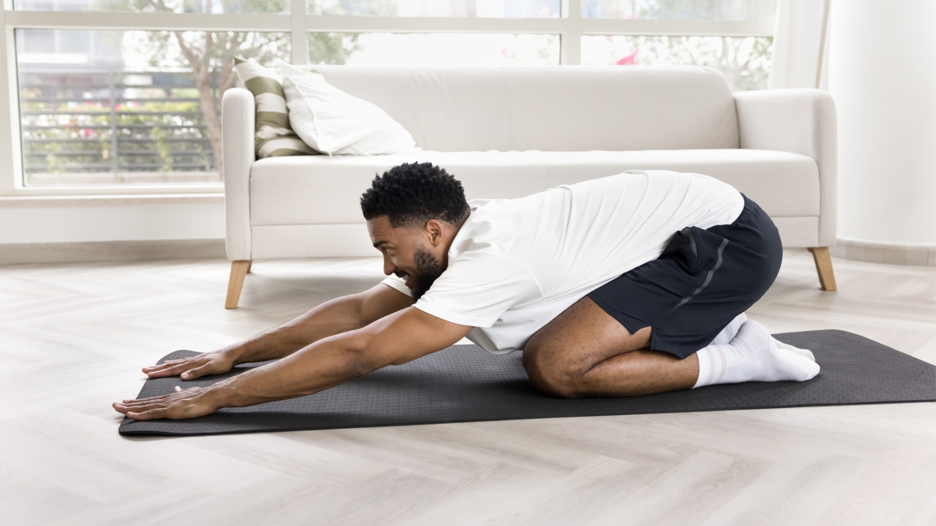 man in a white tshirt and black shorts in a child&#039;s pose position on a black yoga mat with a white sofa behind him. 