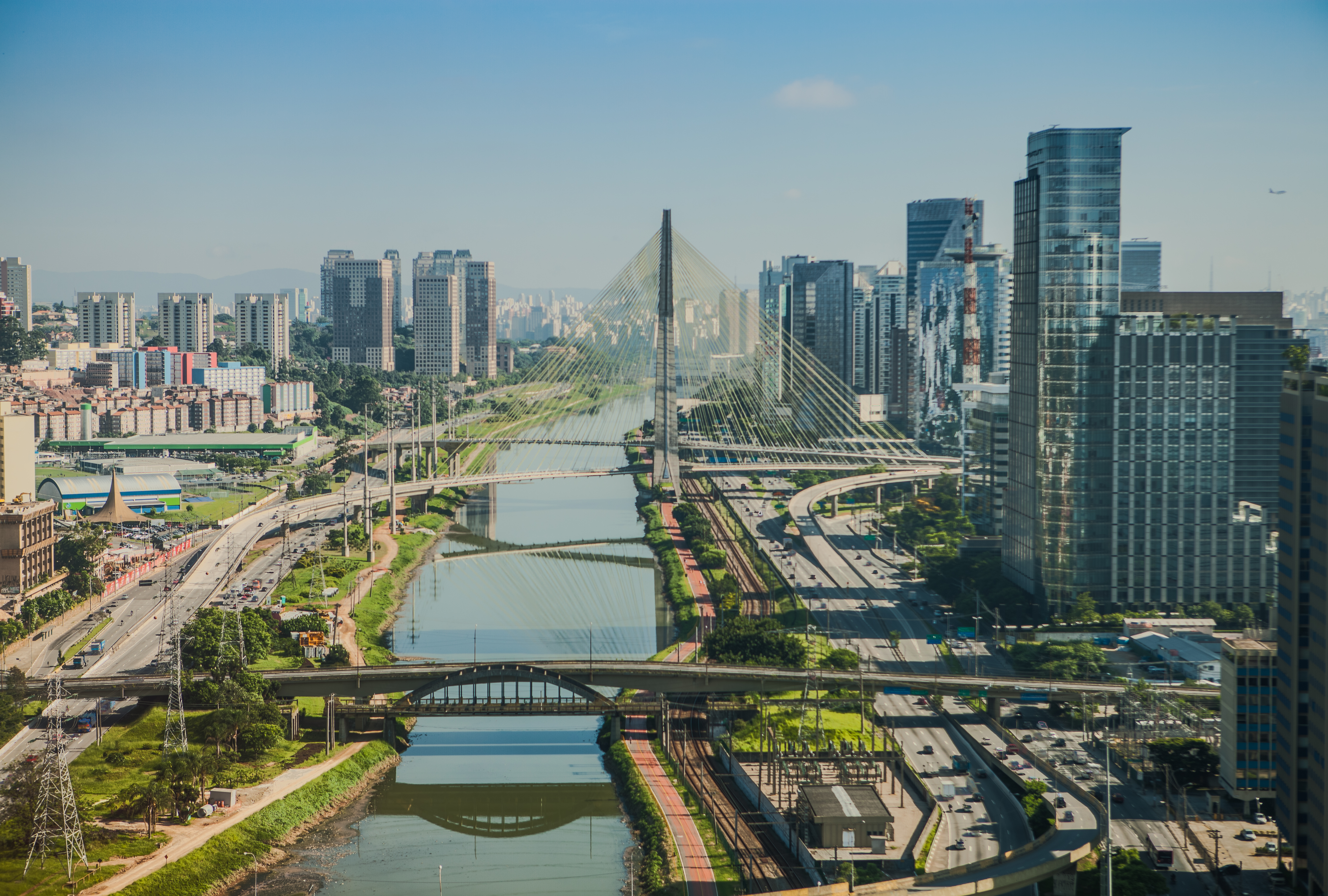 An aerial view of S&amp;atilde;o Paulo in the sunshine, featuring streets, towering bridges, greenery, and skyscrapers.