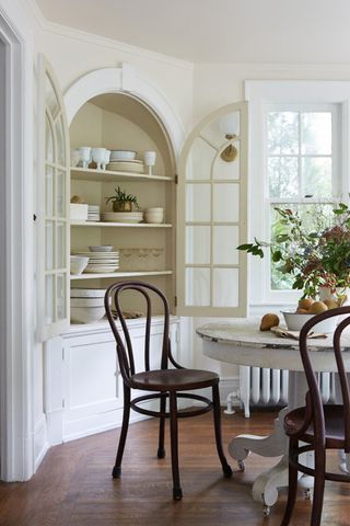 White dining area with rustic farmhouse table and chairs and a white curved display cabinet with doors open