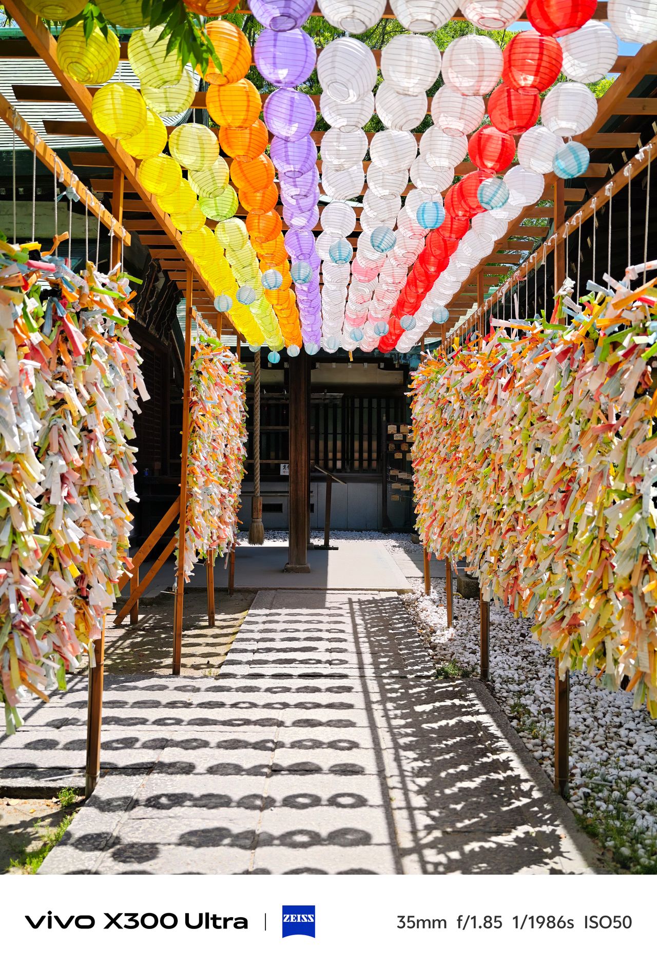 A stone path sheltered by a wooden arbor of colorful lanterns, flanked by walls of tied paper strips.