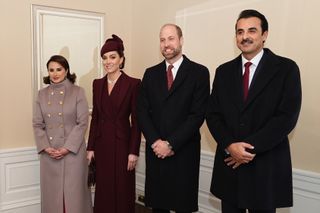 Prince William, Prince of Wales and Catherine, Princess of Wales posing with the Amir of Qatar Sheikh Tamim bin Hamad Al Thani and his wife Sheikha Jawaher, all bundled up in coats and standing with their arms in front of themselves in front of a beige wall.