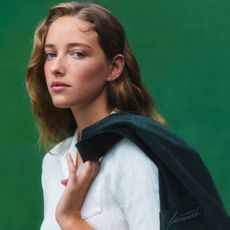 Young blonde woman looking at camera, wearing white Lacoste polo and holding dark green Lacoste blazer over her shoulder, green background.