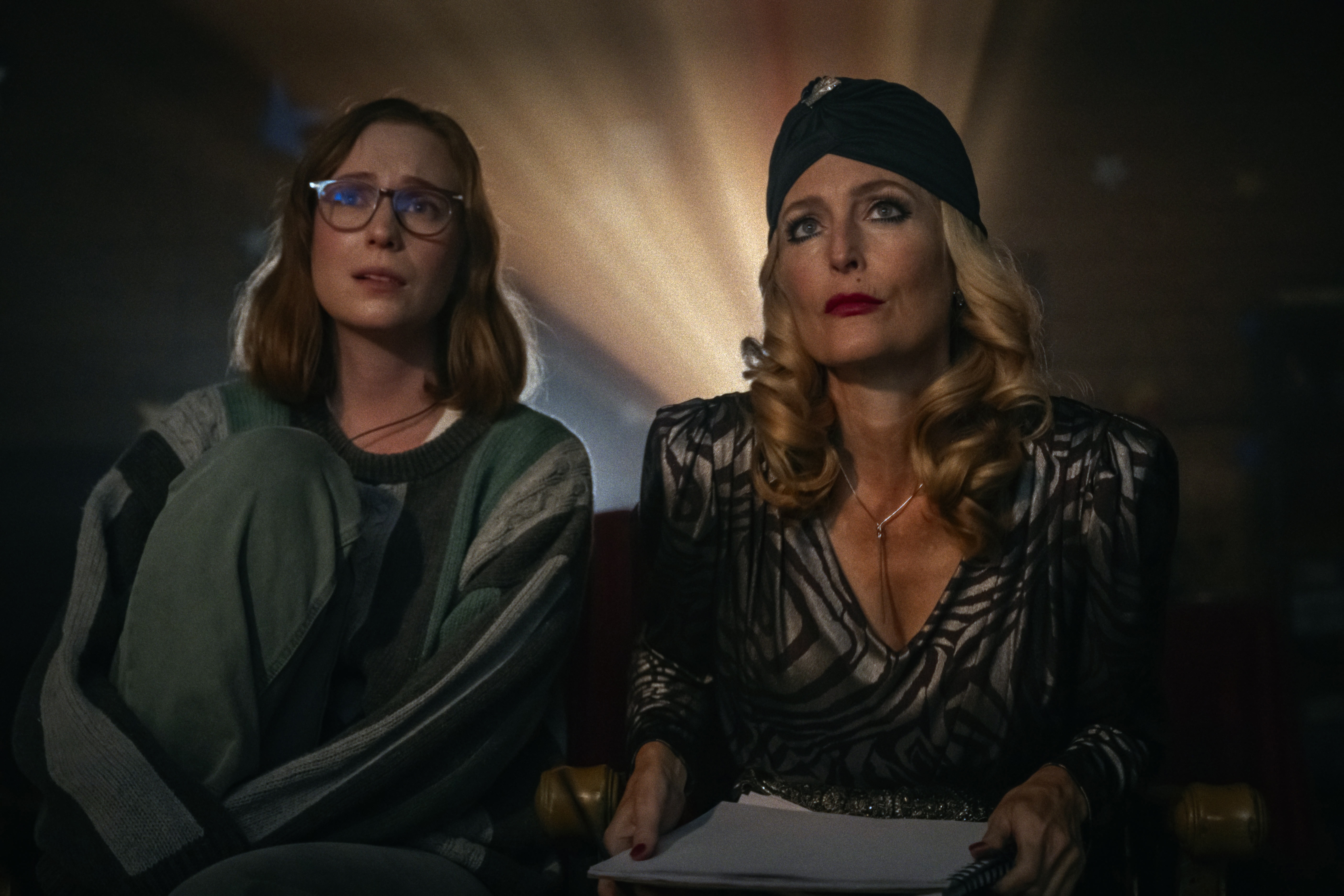 Hannah Einbinder as Kris sits with gillian anderson as billy in an Old Hollywoood-style hair piece and gown in front of the light from a theater projector in 'Teenage Sex and Death at Camp Miasma'