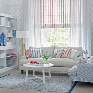 Neutral living room with a cream soda , Red triped Roman blinds and voile curtains and bookshelf againts the wall and a pale blue armchair