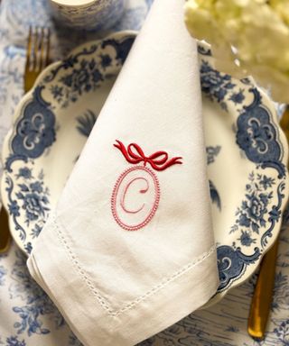 A close-up shot of a white folded napkin with a C embroidered with a bow, and a blue and white plate and a gold knife and fork behind it