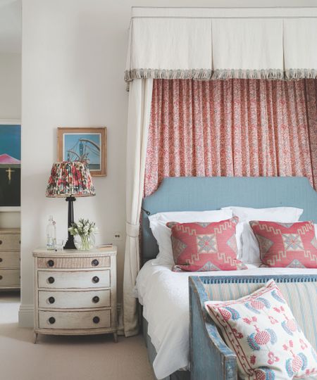A white bedroom with a blue bed with a pink and white canopy, white bedding and patterned cushions. At the end of the bed is a distressed blue bench with striped padding and a patterned cushion, and to the left is a white chest of drawers, with a black lamp with patterned lampshade on top. 
