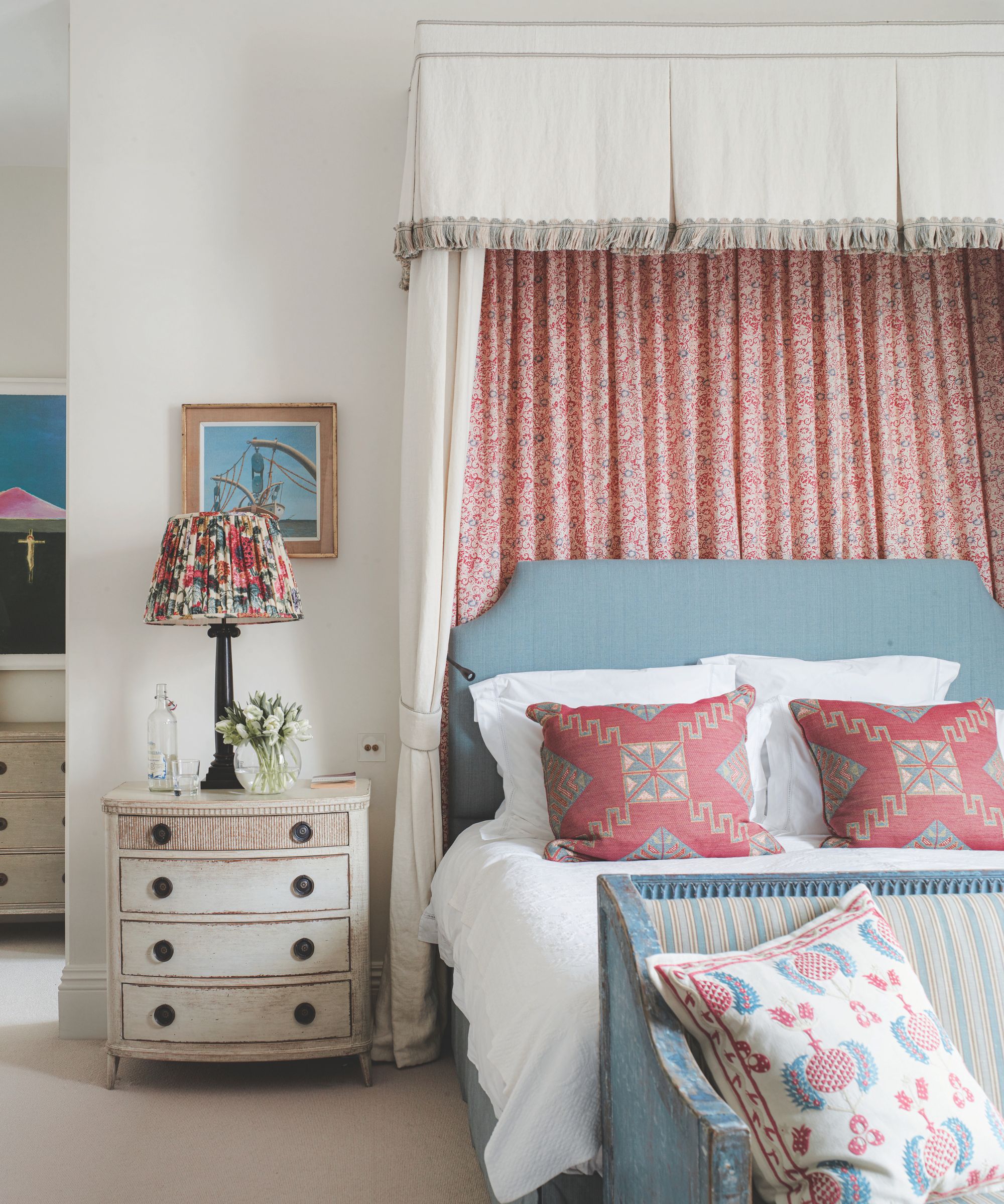 A white bedroom with a blue bed with a pink and white canopy, white bedding and patterned cushions. At the end of the bed is a distressed blue bench with striped padding and a patterned cushion, and to the left is a white chest of drawers, with a black lamp with patterned lampshade on top.
