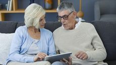 An older couple look at a tablet together while sitting on their sofa.