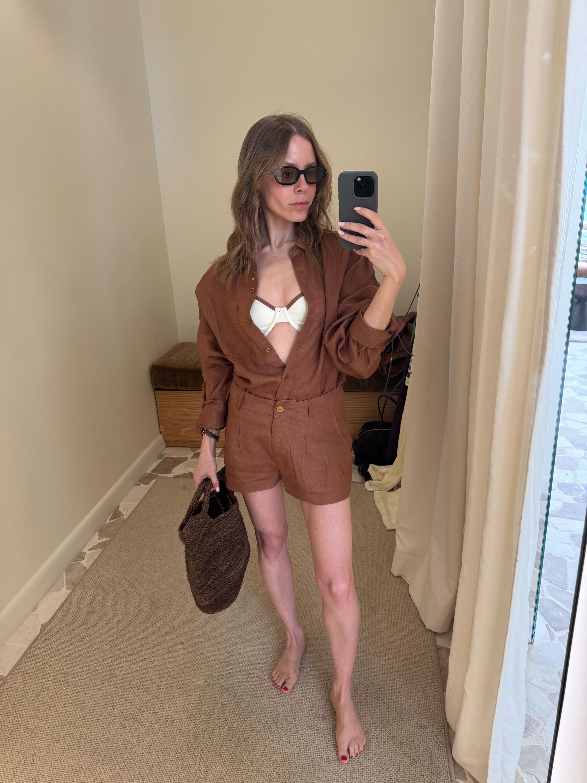 Nikki wears a brown button down shirt and shorts with a white bikini top and brown straw bag.