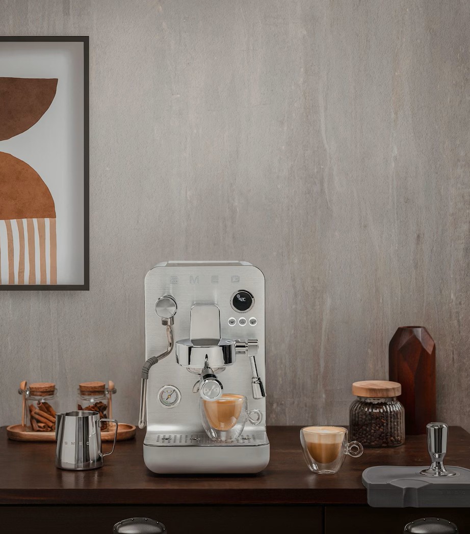 Image of a silver Smeg espresso machine on a dark wooden countertop. The walls are a gray, industrial material. There are a few different coffee mugs and coffee making items scattered on the counter.