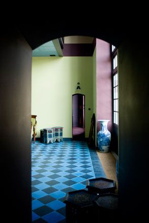 Dark hallway door open leading to another room, blue and black checked floor, pastel coloured walls, blue and white vase under a window, three small side tables, long floor standing mirror