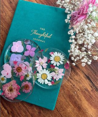 A bird's eye view of a teal blue diary on a wooden table with three pressed flower coasters and white and pink flowers next to them.