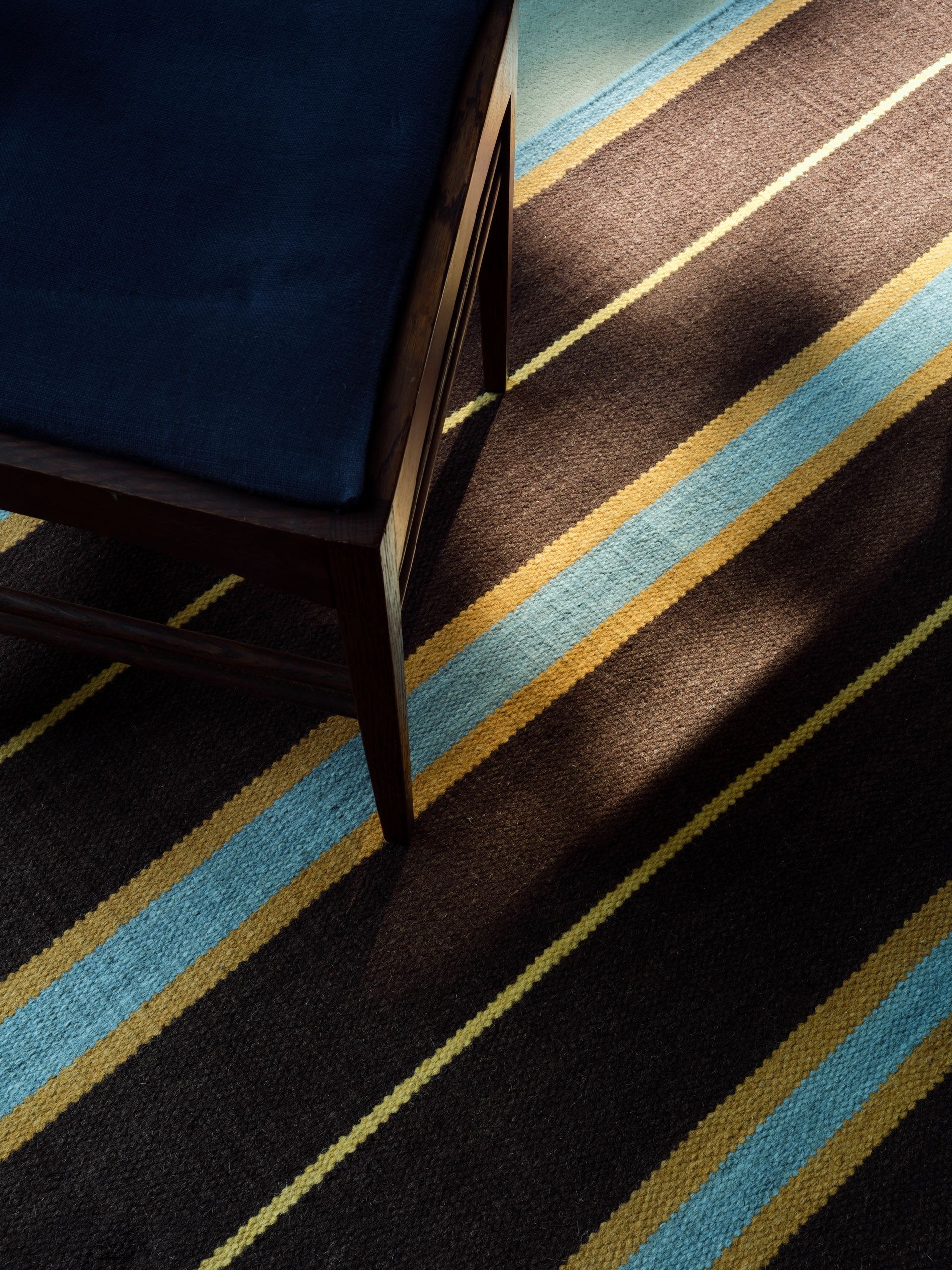 Close up of a brown, yellow, and blue striped rug from above with the corner of a chair in view.